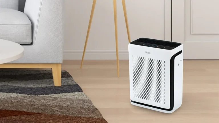 White Levoit Vital 100S-P standing on a wooden floor beside a grey armchair and a floor lamp, showing its size relative to the furniture