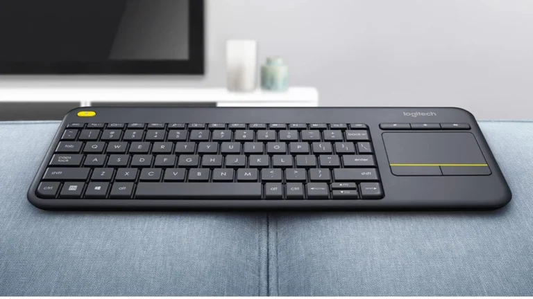 Logitech K400 Plus resting on a grey sofa arm, showing its flat black plastic body, yellow left click button and touchpad