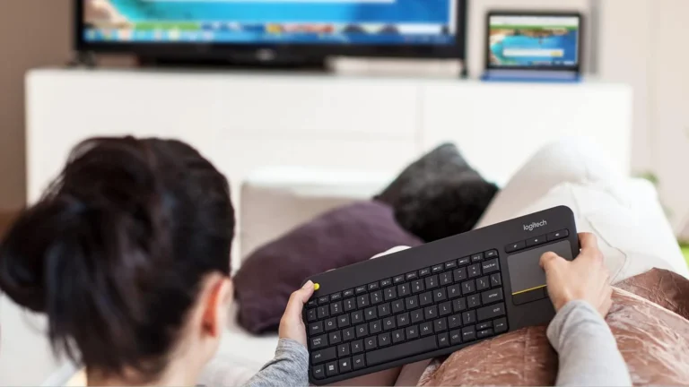 Person on a sofa holding the Logitech K400 Plus on their lap with a thumb near the yellow left click button and a hand on the touchpad, a television in the background